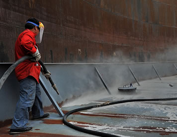 A worker is spraying sand to the bottom surface of ship with sandblast hose.
